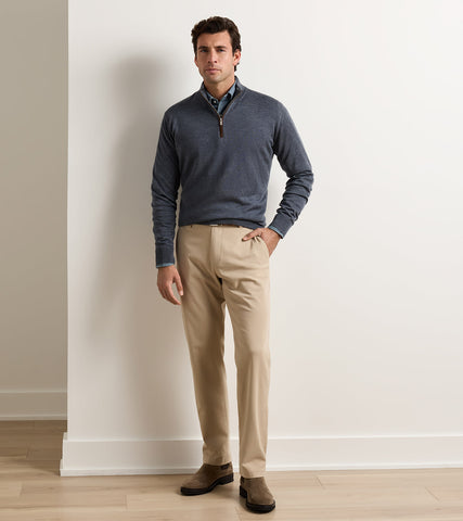 A man modeling a slate blue quarter-zip sweater over a collared shirt and beige trousers, standing against a white wall on a light wood floor.