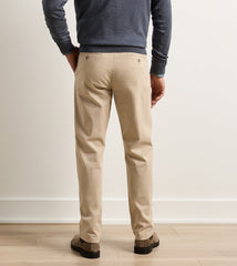 A man viewed from the back wearing beige chino pants, a dark heather gray long-sleeve shirt, and brown shoes, standing on a light wood floor against a white wall.