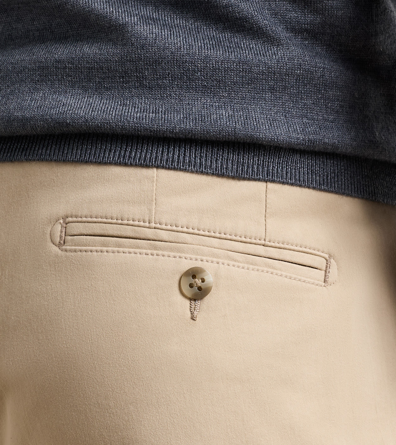 Close-up of the back waistband of beige trousers, showing a buttoned welt pocket and a dark heather gray ribbed knit top folded over the hem.