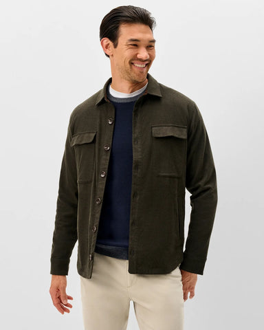 A man wearing an olive green button-up overshirt over a navy sweater and cream pants, photographed against a light gray studio background.
