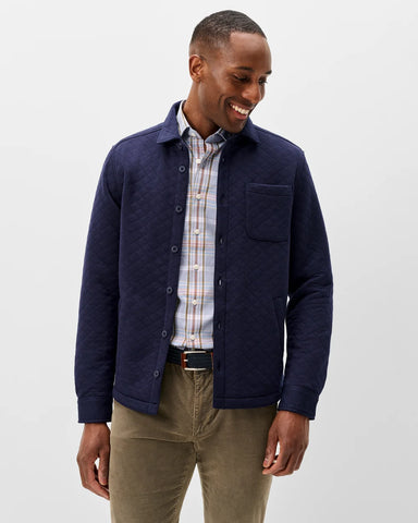 A man wearing a navy blue quilted button-up overshirt over a plaid button-down shirt and khaki pants, standing against a light gray studio background.