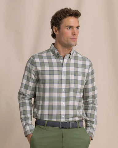 A man wearing a long-sleeve button-down shirt with a green and white checkered pattern, paired with olive green trousers and a dark patterned belt, standing against a neutral beige background.