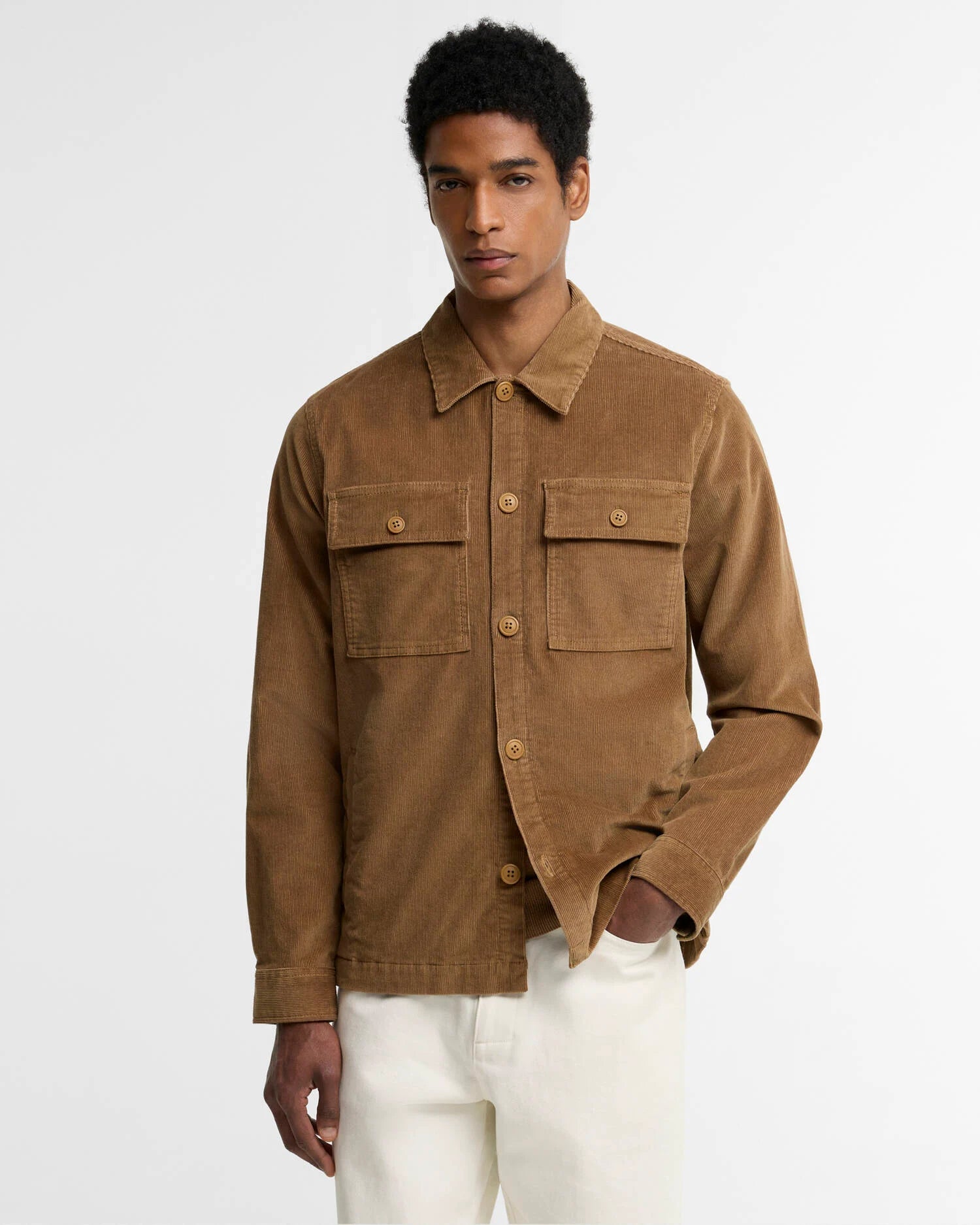 A man wearing a brown corduroy utility jacket with buttoned chest pockets and white pants, standing against a plain light gray studio background.