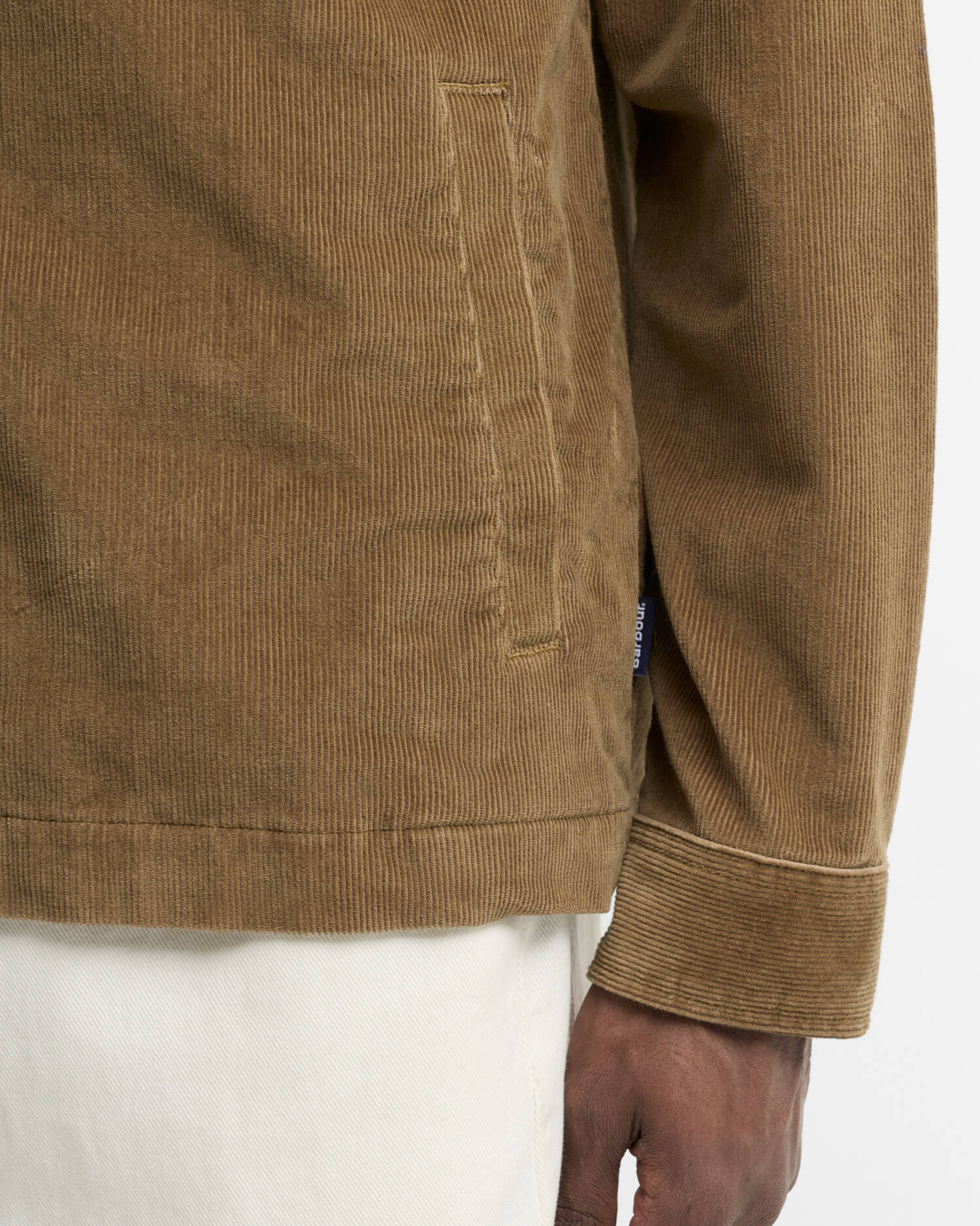 Close-up of a person wearing a brown corduroy jacket, showing the sleeve, pocket, cuff, and part of the hand against a plain light background.
