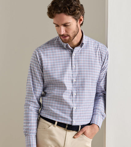 A man wearing a long-sleeve button-down shirt with a fine blue, purple, and white plaid pattern, paired with beige trousers and a black belt, standing against a neutral beige wall.