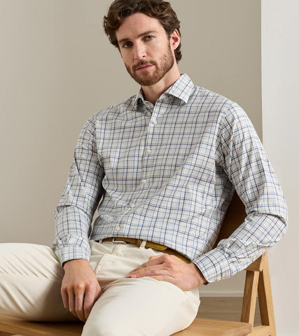 A man wearing a long-sleeve button-down shirt with a fine blue and white windowpane plaid pattern, paired with cream trousers, sitting on a wooden bench against a neutral beige background.