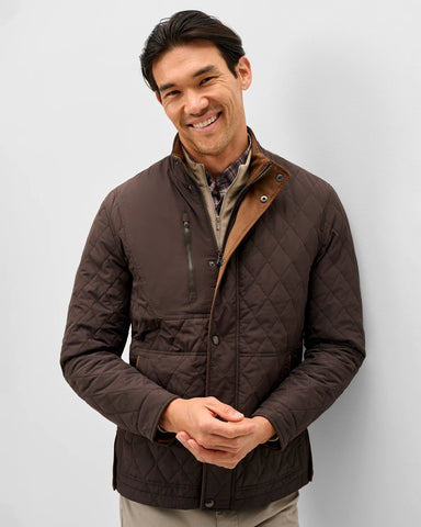 A smiling man wearing a dark brown diamond-quilted jacket with a tan collar lining, standing against a light gray studio background.