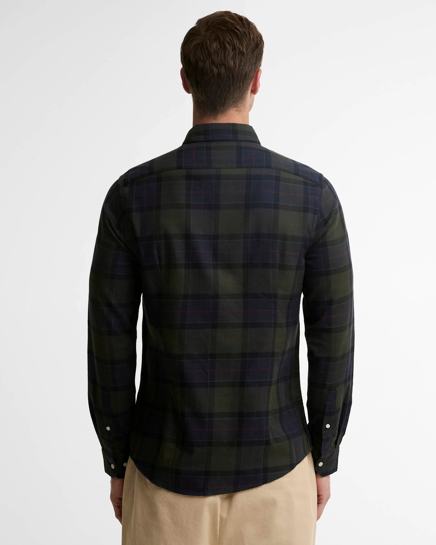 A man viewed from the back wearing a dark green and navy plaid long-sleeve button-up shirt, paired with beige trousers, against a plain white studio background.