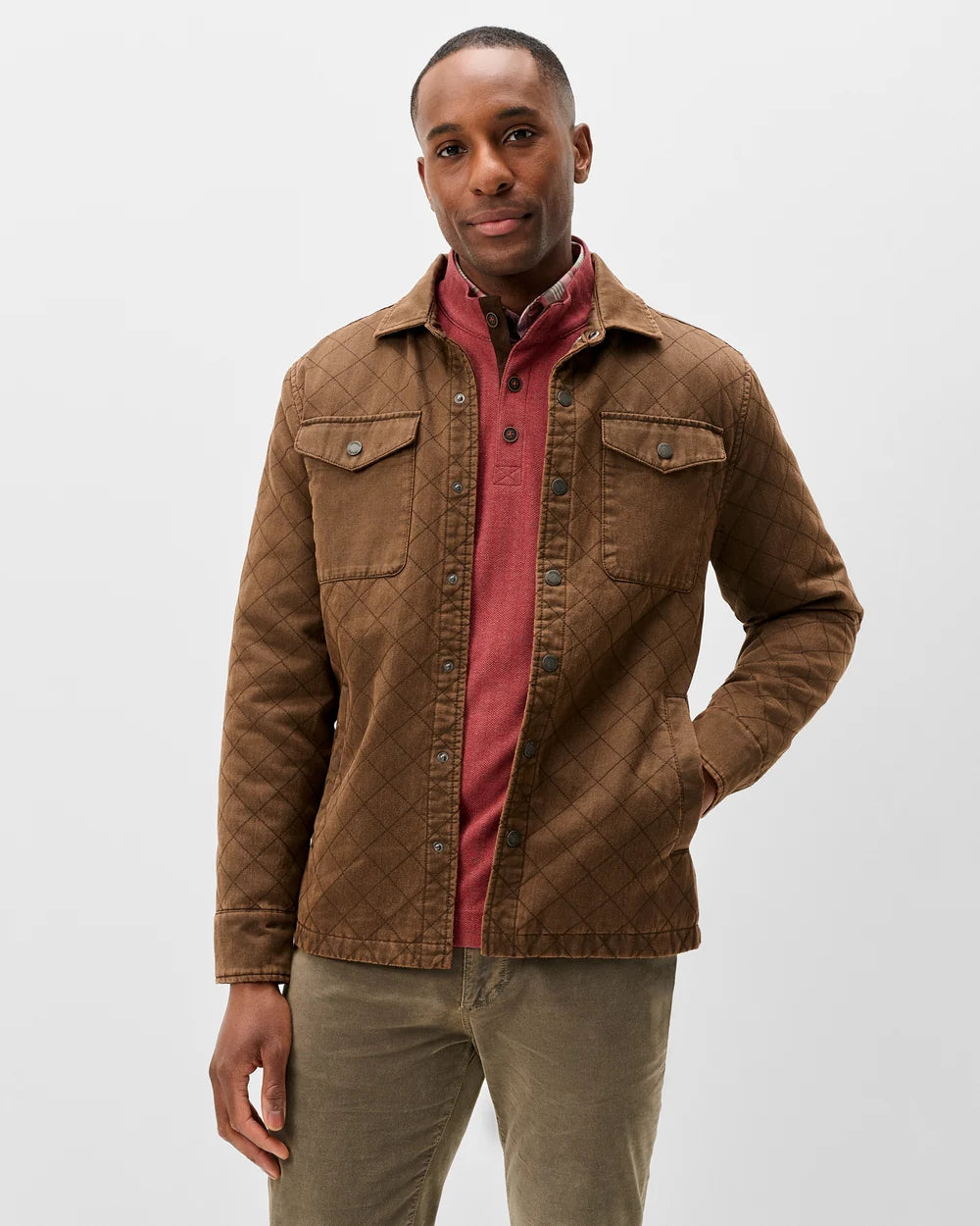 A man wearing a brown quilted overshirt jacket over a dusty red collared shirt and olive green trousers, standing against a light gray studio background.