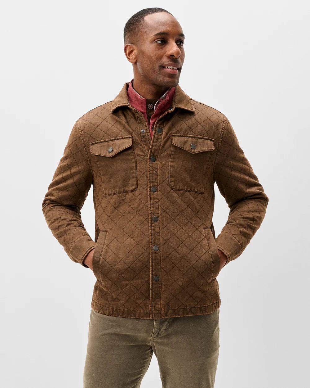 A man wearing a brown diamond-quilted corduroy jacket over a red collared shirt and olive pants, standing against a light gray studio background.