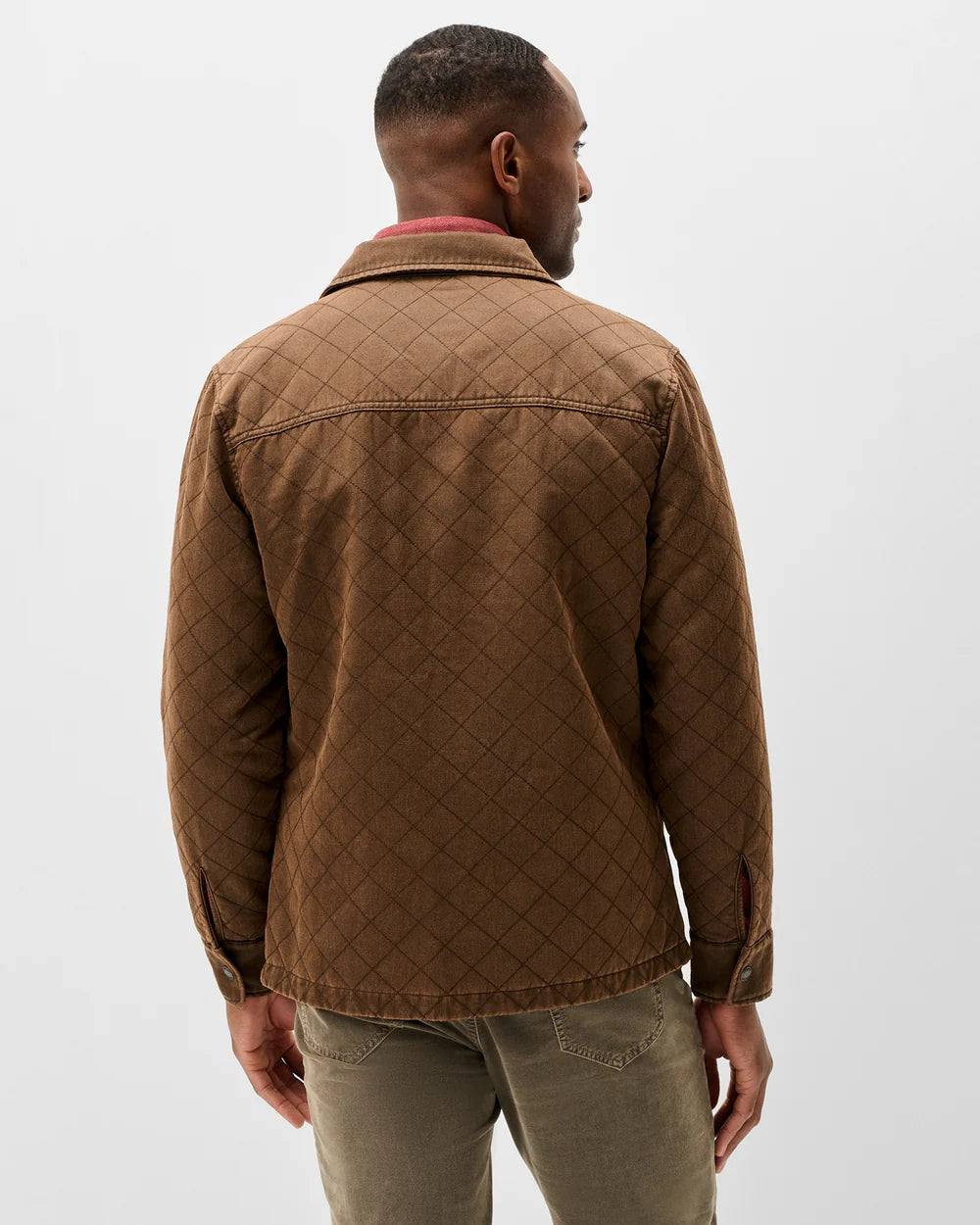 A man viewed from the back wearing a brown quilted jacket and olive green pants against a plain light gray studio background.