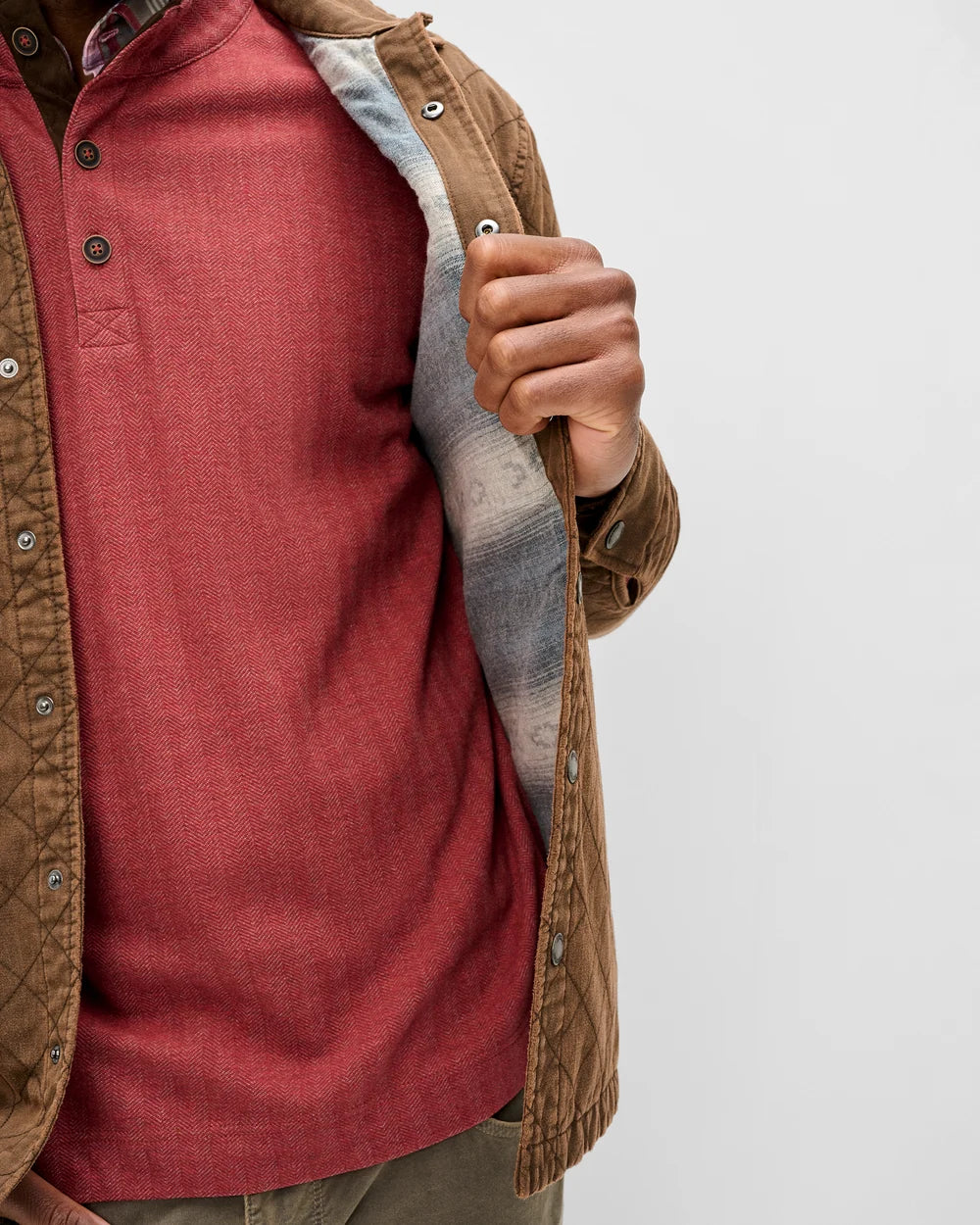 A close-up of a person wearing a brown quilted overshirt over a rust-red henley shirt, holding the collar open to show a gray plaid lining, against a plain light gray background.