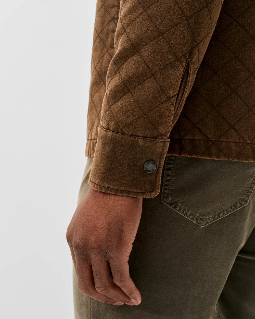Close-up of a person wearing a brown quilted jacket with a buttoned cuff and olive green pants against a light gray studio background.