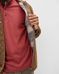 A close-up of a person wearing a brown quilted overshirt over a rust-red henley shirt, holding the collar open to show a gray plaid lining, against a plain light gray background.